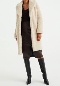 Beige faux fur-kappa med slagkrage, långa ärmar och sidofickor, parat med en mörk, figurnära kjol och knähöga svarta stövlar.