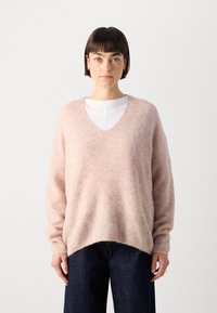 Hellrosa, übergroßer Pullover aus weichem, strukturiertem Stoff mit tiefem V-Ausschnitt und gerippten Bündchen, kombiniert mit dunklen Jeans.