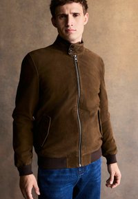 Ruskea mokkanahkainen bomber-takki pystypäällä, edessä vetoketju, sivutaskut ja resorimansetit. Yhdistetty sinisiin farkkuihin.