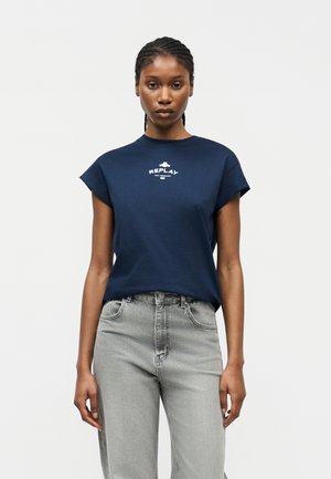 Γυναίκα με πλεξούδα που φοράει αμάνικο μπλε σκούρο T-shirt με το λογότυπο «REPLAY» και ανοιχτό γκρι ψηλόμεσο τζιν, στέκεται μπροστά σε λευκό φόντο.