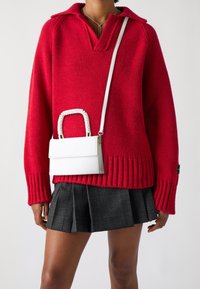 Czerwony sweter dziergany z dekoltem w serek i ściągaczem u dołu, zestawiony z białą torebką o plecionym uchwycie i prostokątnym kształcie, noszony z plisowaną spódnicą w kratkę.