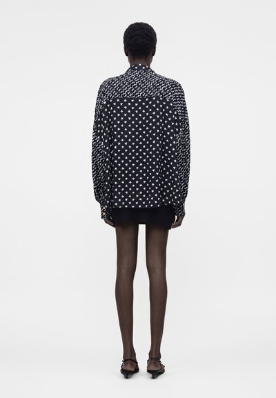 Άτομο που φοράει μαύρο-άσπρο πουκάμισο με αστέρια και ζιγκ-ζαγκ μοτίβα, μαύρη μίνι φούστα, διάφανα καλσόν και μαύρα παπούτσια με χαμηλό τακούνι, στέκεται.