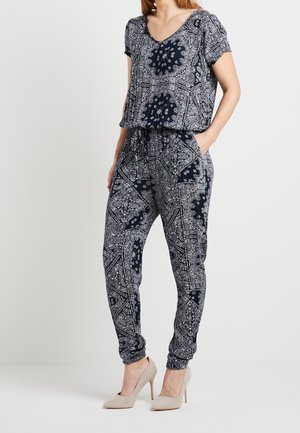 Kobieta ubrana w granatowy kombinezon w biały wzór paisley z krótkimi rękawami i beżowe buty na wysokim obcasie, stojąca z rękami w kieszeniach.