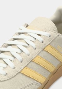 Pantof sport bej din piele întoarsă, cu șireturi albe, trei dungi galbene, partea superioară texturată și talpă din cauciuc lipicioasă. Prezintă un logo galben pe lateral.