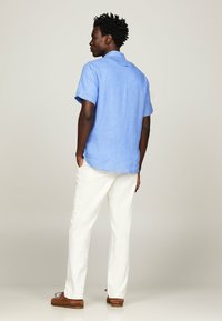 Camicia azzurra chiara, a maniche corte, con bottoni, in tessuto di lino. Abbinata a pantaloni bianchi con gamba dritta e scarpe marroni. Colletto semplice e dettagli sul retro.