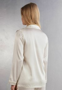 Silkig krämfärgad pyjamasöverkant med kraag med inskärning, långa ärmar och en mjuk textur. Har en delikat söm längs fållen och ärmsluten.