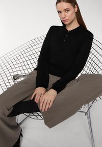 Czarna polo z długim rękawem i trzema guzikami, w połączeniu z szerokimi beżowymi spodniami, siedząca na metalowym krześle z gładką powierzchnią.