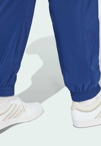 Blå atletiske bukser med elastiske manchetter, parret med hvide sneakers med grå accenter og strukturerede detaljer. Baggrunden er lys.