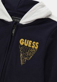 Sudadera con cremallera de color azul marino, con forro blanco en la capucha y texto amarillo de "GUESS" en un patrón triangular en el pecho.