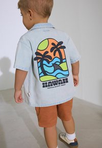 Camisa de manga corta de denim azul claro con un gráfico colorido de playas y palmeras en la parte posterior; combinada con pantalones cortos naranjas y zapatos blancos.