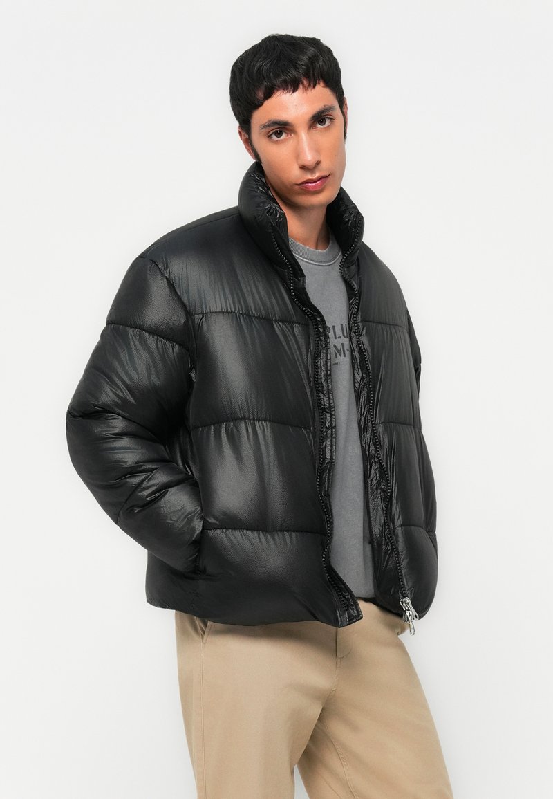 Czarna kurtka puchowa o błyszczącej powierzchni, z zamkiem błyskawicznym, wysokim kołnierzem i pikowanym wzorem; noszona na szarym podkoszulku i w połączeniu z jasnymi khaki spodniami.