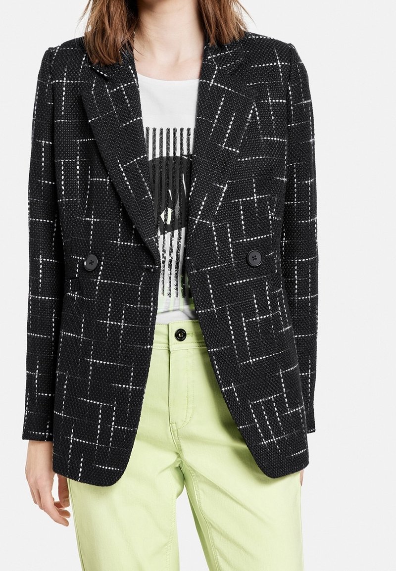 Femme portant un blazer noir texturé avec un motif de grille blanche sur un t-shirt graphique blanc et un pantalon vert clair, debout devant un fond uni.