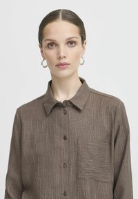 Chemise brune à boutons, en tissu texturé, avec un col, des manches longues et une poche poitrine ornée de légères rayures blanches.