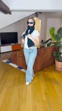 Una donna in una maglietta bianca e jeans blu chiaro, con occhiali da sole e una sciarpa a quadri verde scuro, posa in un ambiente moderno di soggiorno.