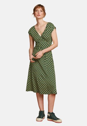 Femme portant une robe portefeuille verte à motif avec des manches courtes et des baskets montantes vert foncé, debout les mains jointes.