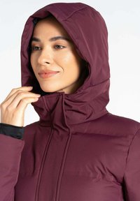 Burgunderfarbene isolierte Jacke mit hohem Kragen und abnehmbarer Kapuze. Glatte Außenstruktur, mit Reißverschluss und Seitentaschen.