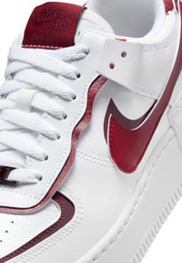 Biały sneaker z teksturowanej skóry, czerwonymi akcentami i wyraźnym logo Nike. Posiada białe sznurowadła i wyściełany język dla komfortu.