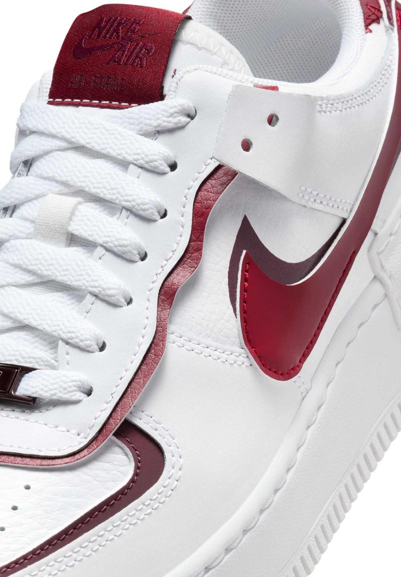 Hvid sneaker med struktureret læder, røde accenter og fremtrædende Nike-logo. Har hvide snørebånd og en polstret tunge for komfort.