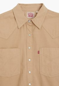 Camicia in corduroy beige con bottoni, caratterizzata da yoke appuntiti, due tasche sul petto, bottoni argentati e un'etichetta rossa sulla tasca sinistra.