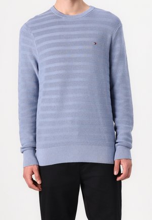 Homme portant un pull en tricot texturé bleu clair avec des rayures horizontales et un petit logo sur la poitrine, associé à un pantalon noir.