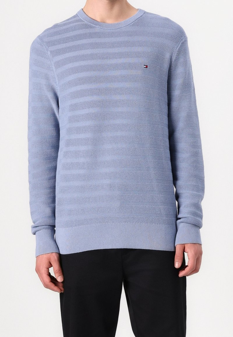 Homme portant un pull en tricot texturé bleu clair avec des rayures horizontales et un petit logo sur la poitrine, associé à un pantalon noir.