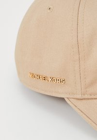 Beige baseballcap laget av stoff, med en buet skjerm og en gull "MICHAEL KORS" logo på siden. Teksturert, uformelt design.