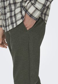 Groene broek met een herringbone-patroon en een getailleerde pasvorm, gecombineerd met een grijs- en zwartgeruite flanellen overhemd, waarbij de knoopdetails van het overhemd zichtbaar zijn.