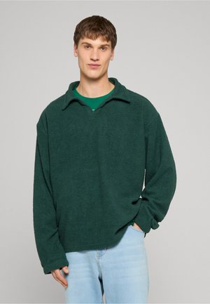 Fiatal férfi rövid barna hajjal, sötétzöld mintás pulóvert és világoskék farmert visel, egyik kezét a zsebében tartva, egyszínű háttér előtt áll.