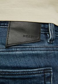 Vaqueros de mezclilla azul oscuro con una etiqueta de cuero negro grabada con "CHASIN'" en la parte trasera de la cinturilla, con sutil desgaste y detalles de costura.