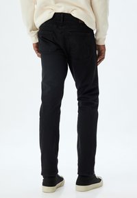 Jeans de mezclilla negra mostrados desde la parte trasera, con un diseño ajustado y costuras visibles. Dos bolsillos traseros con una sutileza en la marca. Forma de pierna slim.