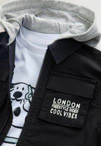 Črna jakna z zadrgo in sivo kapuco, z belim napisom. Pod njo je bela majica s potiskom risanega psa v črni obris.