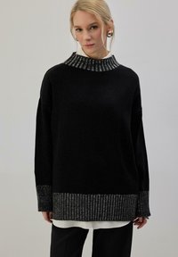 Pull noir avec un col montant à motifs et des accents métalliques. Comprend des manches longues et un ourlet texturé. Porté sur un haut blanc.