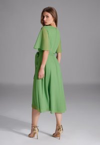 Robe midi verte fluide avec des manches courtes en voile, une taille nouée et un détail en trou de serrure à l'arrière. Associée à des sandales à talons dorées.