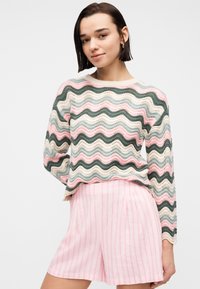 Νεαρή γυναίκα με κοντά σκουρόχρωμα μαλλιά, φορώντας πουλόβερ με πολύχρωμο κυματιστό σχέδιο και ροζ ριγέ σορτς, ποζάροντας με το ένα χέρι πίσω από την πλάτη της.