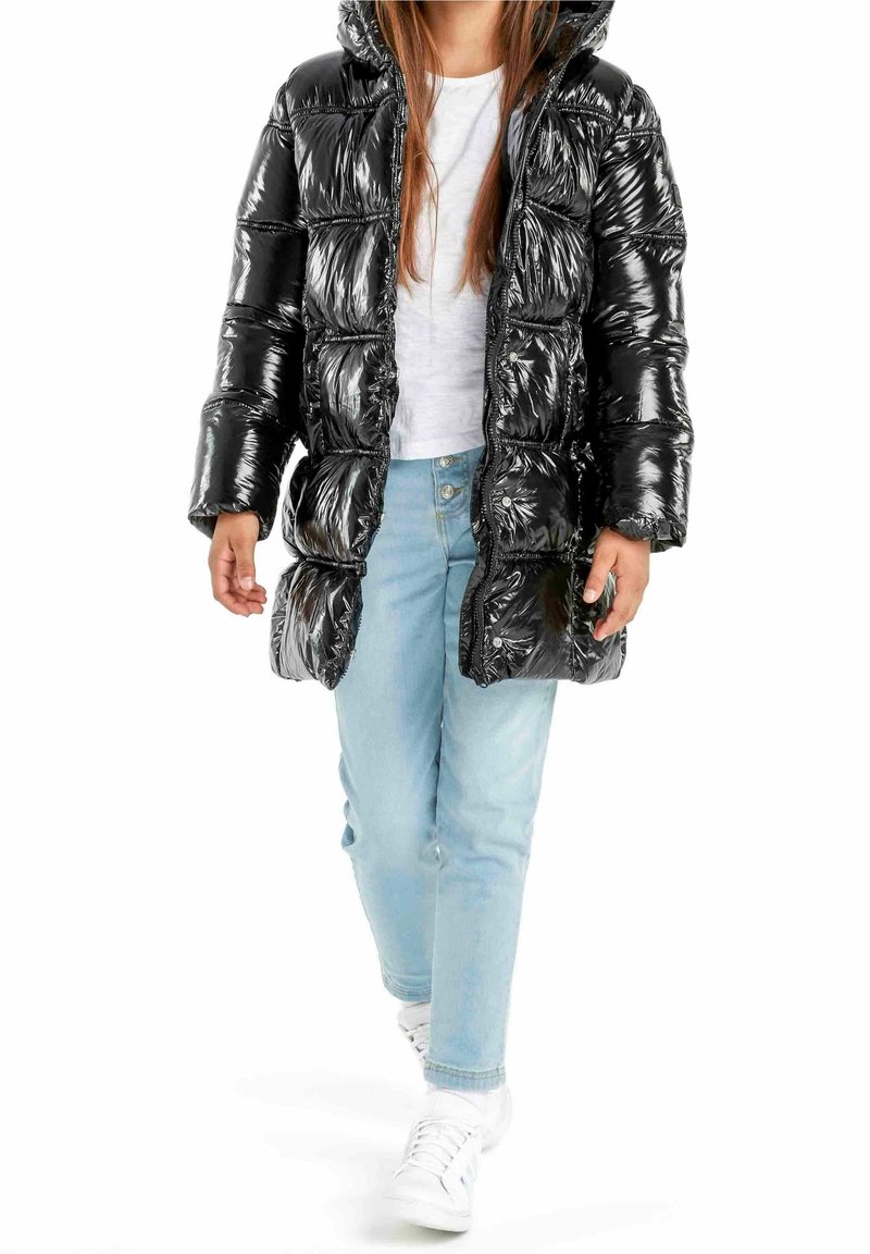 Enfant portant un manteau matelassé noir brillant par-dessus une chemise blanche et un jean bleu clair, marchant vers l'avant avec des baskets blanches.