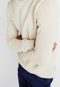 Krämfärgad stickad tröja med texturmönster, ribbade manschetter och hög krage, i kombination med marinblå byxor.