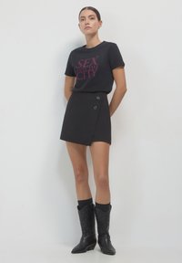 Frau trägt ein schwarzes "Sex and the City" T-Shirt, einen schwarzen asymmetrischen Rock mit Knöpfen und schwarze bestickte Cowboystiefel, steht vor einem weißen Hintergrund.