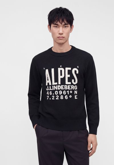 Jeune homme portant un pull noir avec un texte blanc indiquant "ALPES J. LINDEBERG 46.0961° N 7.2286° E", debout devant un fond uni.