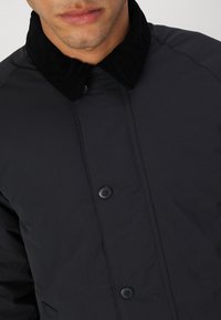 Casaco preto feito de tecido suave com colarinho de veludo cotelê preto, apresentando frente com botões e bolsos laterais. Design simples e funcional.