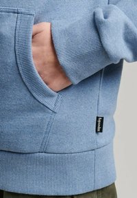 Sudadera azul claro con un bolsillo canguro, puños y dobladillo acanalados. Presenta una pequeña etiqueta negra en el lateral con el nombre de la marca.