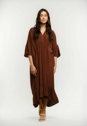 Młoda kobieta stoi w luźnej, długiej brązowej sukience z marszczeniami, nosząc beżowe sandały na otwarty palec, na tle jednolitego jasnego tła.