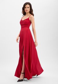 Rode jurk tot op de vloer met een gedrapeerde buste, dunne bandjes en een hoge split tot op de dij. Gemaakt van zachte stof met een lichte glans. Zwarte hoge hakken.