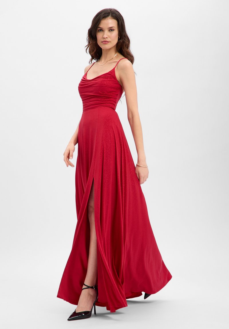 Rode jurk tot op de vloer met een gedrapeerde buste, dunne bandjes en een hoge split tot op de dij. Gemaakt van zachte stof met een lichte glans. Zwarte hoge hakken.