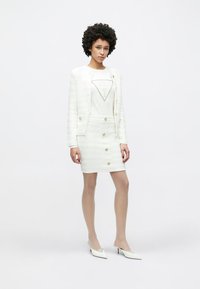 Λευκό σακάκι προσαρμοσμένης ραφής με χρυσά κουμπιά και υφή, σε συνδυασμό με φούστα που εφαρμόζει και φέρει παρόμοια κουμπιά, και ένα γραφικό μπλουζάκι από κάτω.