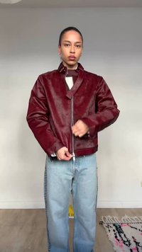 Άτομο με μαλλιά δεμένα πίσω, φορώντας κόκκινο δερμάτινο μπουφάν και ανοιχτό μπλε τζιν, κλείνει το φερμουάρ του μπουφάν ενάντια σε απλό φόντο τοίχου.