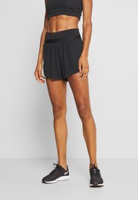 Svart idrottsbyxor med bred midja, strukturerad tyg och veckad design, tillsammans med svarta löparskor.