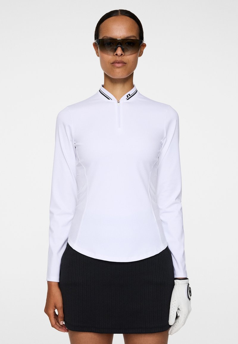 Haut de golf blanc à manches longues avec un col à demi-fermeture éclair, des panneaux latéraux noirs, et un logo sur le col ; associé à une jupe plissée noire et un gant blanc.