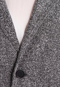 Šedá látka s vzorem houndstooth s texturovaným tkaním, s černým knoflíkem na oblasti límce blazera.
