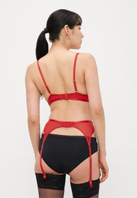 Raudonas liemenėlė su reguliuojamomis petnešėlėmis ir kabliuko užsegimu, derinamas su atitinkančia juostos diržu. Abu turi lygią tekstūrą ir funkcionalią aparatūrą.