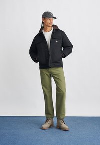 Casaco preto com colarinho castanho, calças verdes, boné cinza e botas beges. Apresenta grandes bolsos frontais e um design de ajuste relaxado.